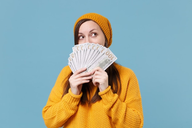 Pensive young woman girl in sweater and hat posing isolated on blue background. People sincere emotions lifestyle concept. Mock up copy space. Covering face with fan of cash money in dollar banknotes.