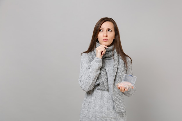 Pensive young woman in gray sweater, scarf looking up, put hand prop up on chin, holding daily pill box isolated on grey background. Healthy lifestyle, ill sick disease treatment, cold season concept.