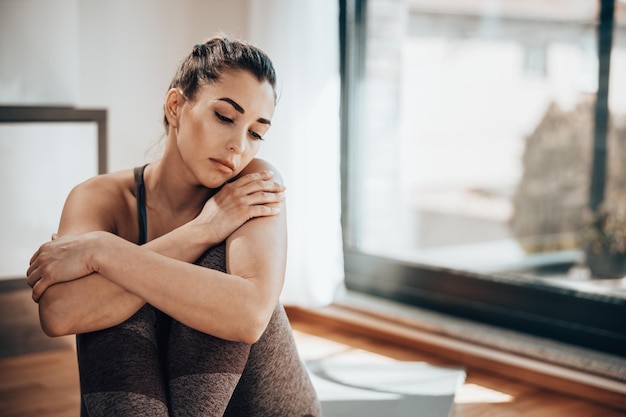 Pensive young woman is sitting on the yoga mat and preparing for training at home.