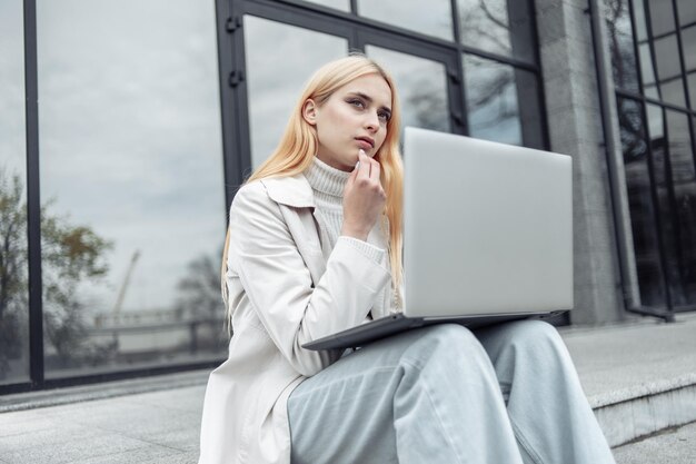 Pensive young woman sitting with laptop on stairs near business center