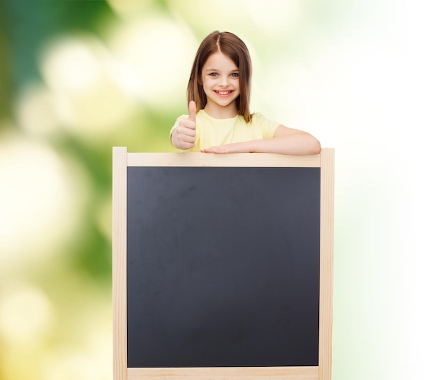 people, advertisement and education concept - happy little girl with blank blackboard showing thumbs up