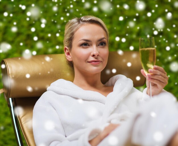 people, beauty, lifestyle, holidays and relaxation concept - beautiful young woman in white bath robe lying on chaise-longue and drinking champagne at spa with snow effect