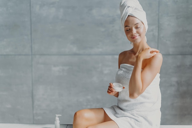 People beauty spa and cosmetology concept Relaxed young European female puts body cream touches shoulder gently wrapped in white soft towel closes eyes with pleasure poses against grey wall