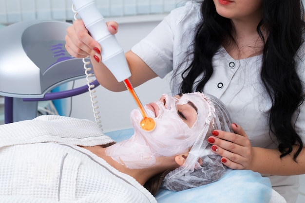 People, beauty, spa, cosmetology and technology concept - close up of beautiful young woman lying with closed eyes having face massage by massager in spa.