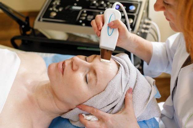 People, beauty, spa, cosmetology and technology concept - The cosmetologist makes the procedure ultrasonic face peeling of the facial skin of a beautiful, young woman in a beauty salon