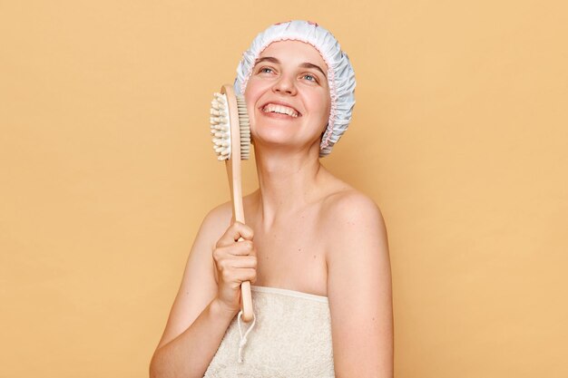 People and body treatment after shower Smiling beautiful young woman using dry brush for body has healthy smooth skin smiles gently standing against beige background wears showr cap