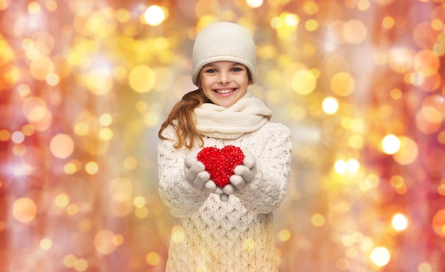 people, charity, holidays, children and love concept - smiling teenage girl in winter clothes with small red heart over lights background