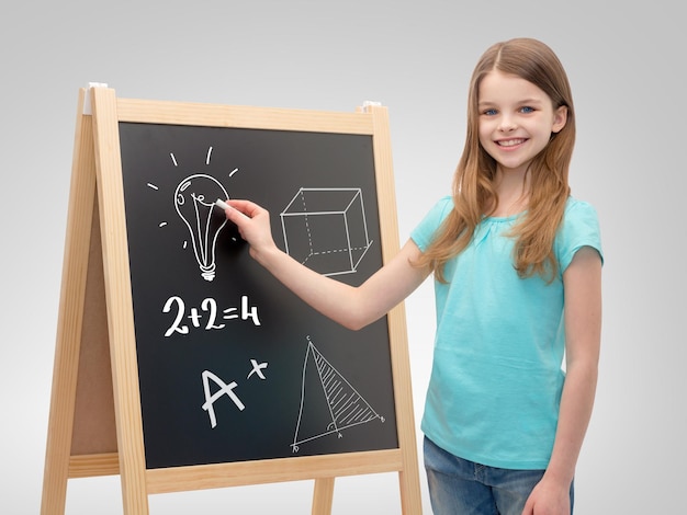 people, childhood and education concept - happy little girl with chalk drawing on blackboard