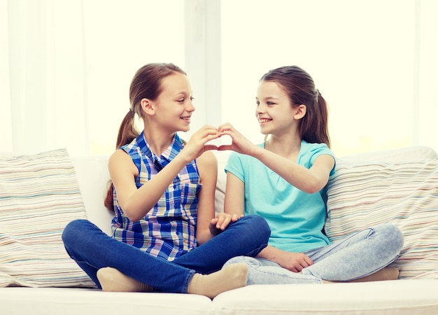 people, children, friends and friendship concept - happy little girls sitting on sofa and showing heart shape hand sign at home