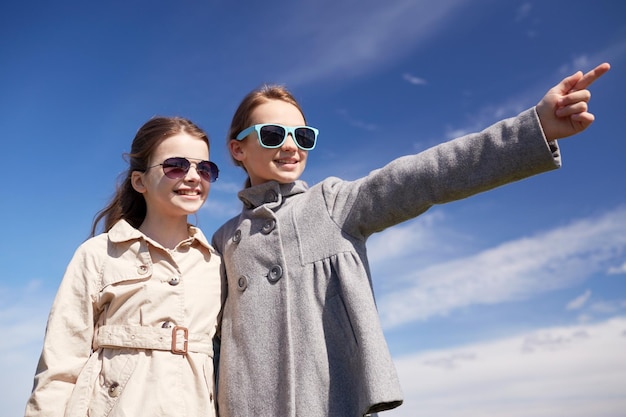 people, children, friends and friendship concept -happy little girls in sunglasses hugging and pointing finger outdoors