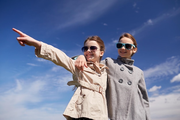 people, children, friends and friendship concept -happy little girls in sunglasses hugging and pointing finger outdoors