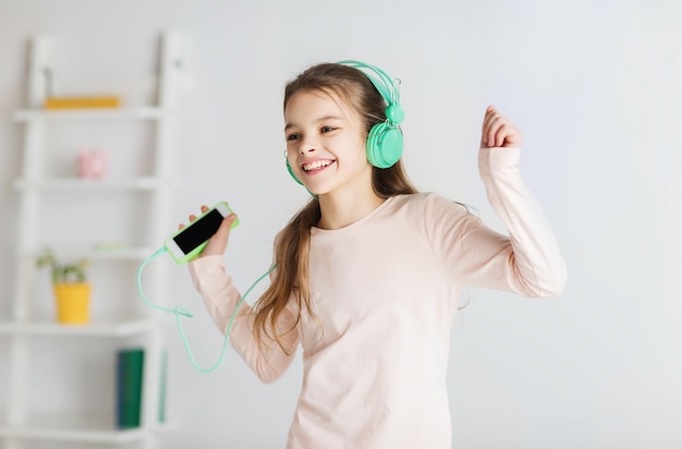 people, children, pajama party and technology concept - happy smiling girl in headphones with smartphone and listening to music at home