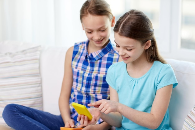 people, children, technology, friends and friendship concept - happy little girls with smartphones sitting on sofa at home