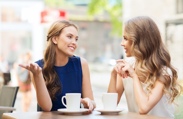 people, communication and friendship concept - smiling young women drinking coffee or tea and talking at outdoor cafe