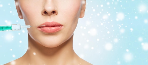 people, cosmetology, plastic surgery and beauty concept - close up of beautiful young woman face and syringe making injection for lips augmentation over blue background and snow