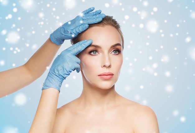 people, cosmetology, plastic surgery and beauty concept - surgeon or beautician hands touching woman face over blue background and snow