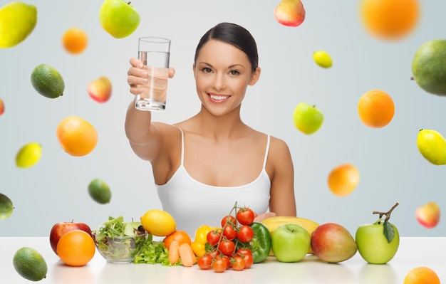 people, diet and vegetarian concept - happy asian woman with healthy food showing glass of water over gray background with falling fruits