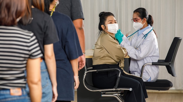 People of different ages in a row standing and waiting for a vaccine injection while doctor giving flu or pneumonia shot to senior woman in face mask. Covid-19 or coronavirus vaccination concept.