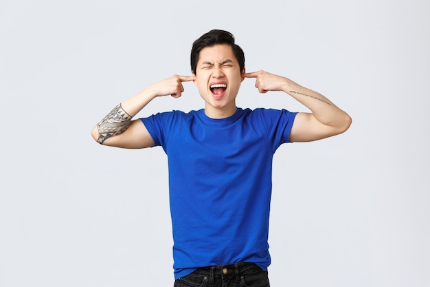 People different emotions, lifestyle and casual concept. Depressed and angry young asian man in blue t-shirt, shut ears and shouting distressed, have breakdown, unwilling to listen, grey background.