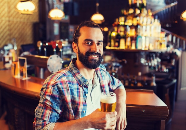 people, drinks, alcohol and leisure concept - happy young man drinking beer at bar or pub