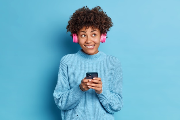 People entertainment and leisure concept. Pleased African American woman bites lips wears stereo headphones listens audio track enjoys favorite melody dressed in sweater