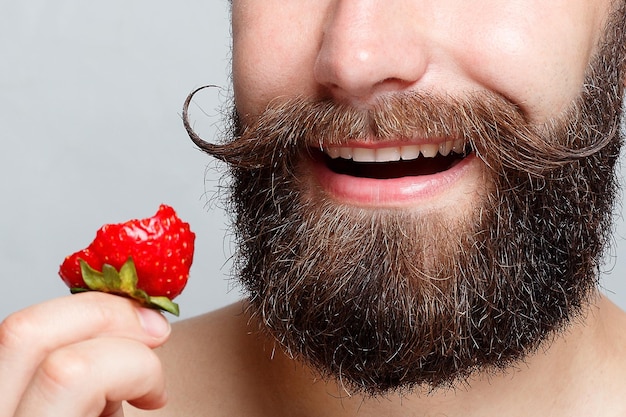 People food beauty lifestyle valentines day concept closeup portrait young man holding a strawberry and smiling Portrait of handsome bearded and mustache on a gray background