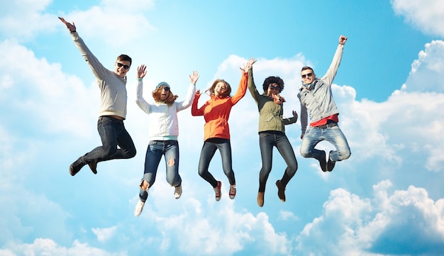 people, freedom, happiness and teenage concept - group of happy friends in sunglasses jumping high over blue sky and clouds background