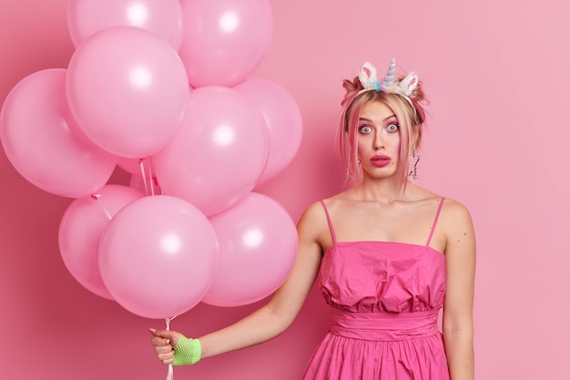 People holidays celebration concept. Shocked beautiful woman wears festive dress unicorn headband celebrates birthday organises party holds bunch of inflated balloons 