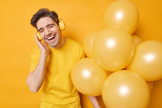 People holidays celebration and fun concept Happy brunet adult man enjoys favorite melody keeps hand on headphones poses with helium balloons dressed casually isolated over yellow background