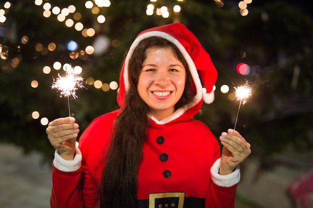 People, holidays and christmas concept - young woman in Santa costume with sparklers or Bengal light