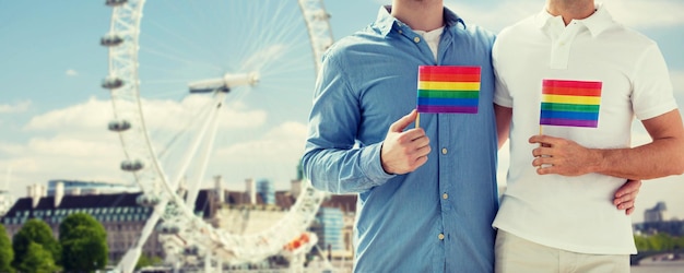 people, homosexuality, same-sex marriage, travel and love concept - close up of happy male gay couple holding rainbow flags and hugging from back over london ferry wheel background