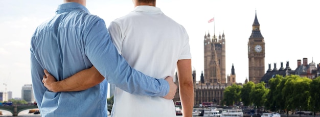 people, homosexuality, same-sex marriage, travel and love concept - close up of happy male gay couple hugging from back over big ben and houses of parliament in london background