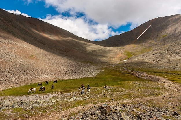 People and horses rest before crossing the pass Horses graze on the grass A path with stones through the mountains Clouds over the mountains Horizontal