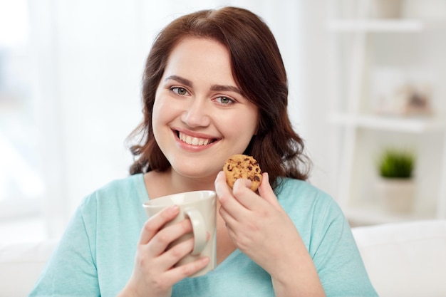 people, junk food, unhealthy eating and leisure concept - happy plus size young woman with cup of tea with cookie at home