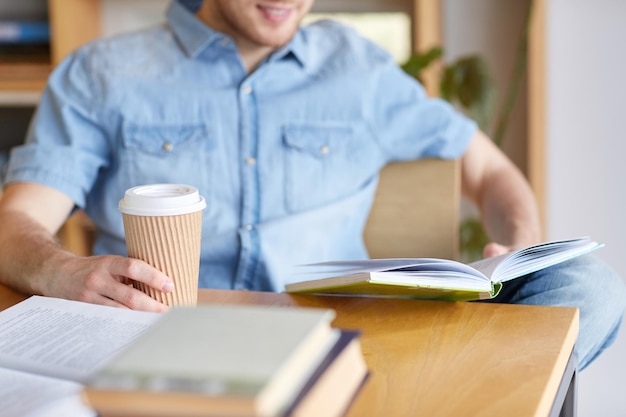 people, knowledge, education and school concept - happy student reading book and drinking coffee in library