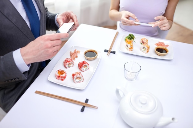 people, leisure, eating, food and technology concept - close up of couple with smartphones taking picture of sushi at restaurant