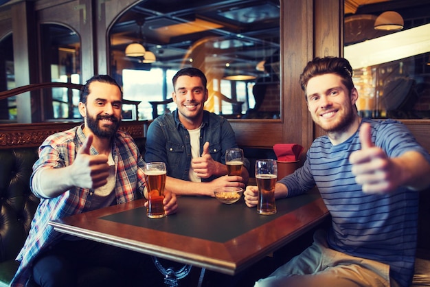 people, leisure, friendship and and bachelor party concept - happy male friends drinking beer and showing thumbs up at bar or pub