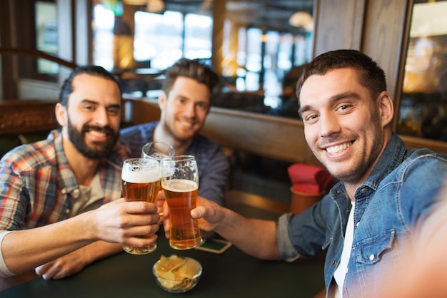 people, leisure, friendship, technology and bachelor party concept - happy male friends taking selfie and drinking beer at bar or pub