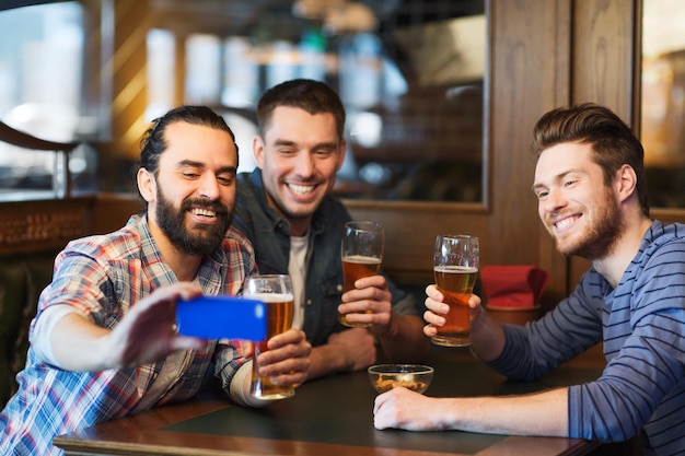 people, leisure, friendship, technology and bachelor party concept - happy male friends with smartphone taking selfie and drinking beer at bar or pub