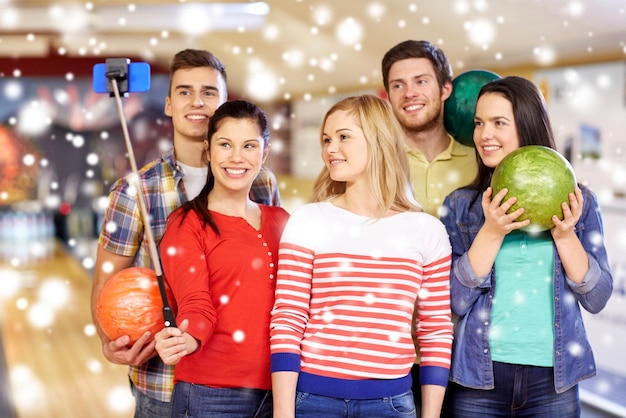 people, leisure, sport, friendship and entertainment concept - happy friends taking selfie with smartphone on monopod in bowling club at winter season