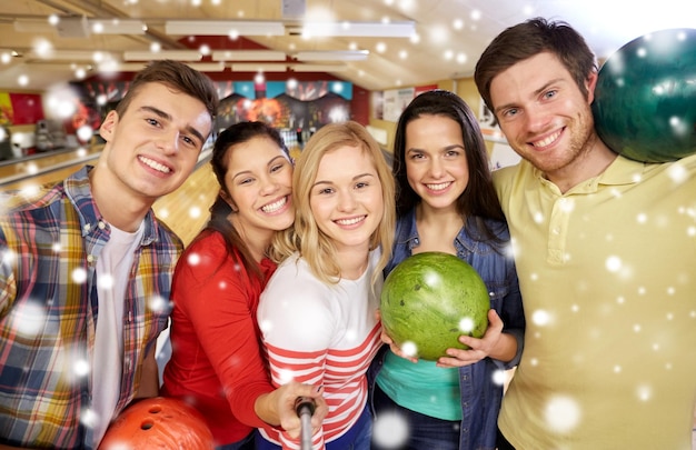 people, leisure, sport, friendship and entertainment concept - happy friends taking selfie with smartphone on monopod in bowling club at winter season