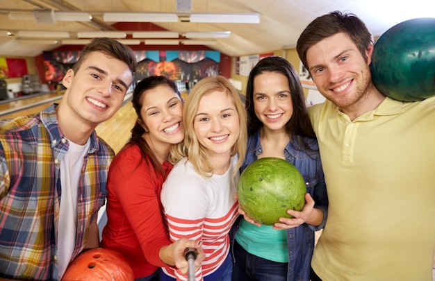 people, leisure, sport, friendship and entertainment concept - happy friends taking selfie with smartphone on monopod in bowling club