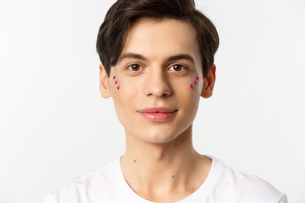 People, lgbtq and beauty concept. Close-up of happy queer guy with applied lip gloss and glitter, smiling and looking at camera, standing over white background.