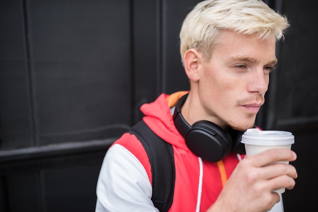 People and lifestyle concept Portrait of handsome Caucasian male with backpack and headphones relaxing during coffee break enjoy a mug of coffee outdoor against dark background in the city