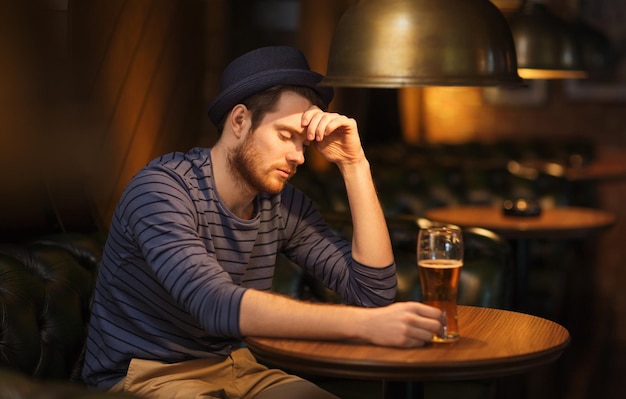 people, loneliness, alcohol and lifestyle concept - unhappy single young man in hat drinking beer at bar or pub