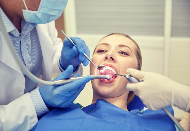 people, medicine, stomatology and health care concept - close up of dentists and assistant with mirror, drill and dental air water gun spray treating female patient teeth at dental clinic