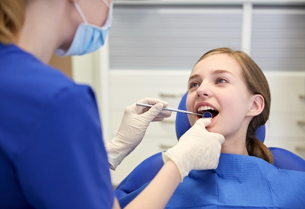 people, medicine, stomatology and health care concept - happy female dentist with mirror checking patient girl teeth up at dental clinic office