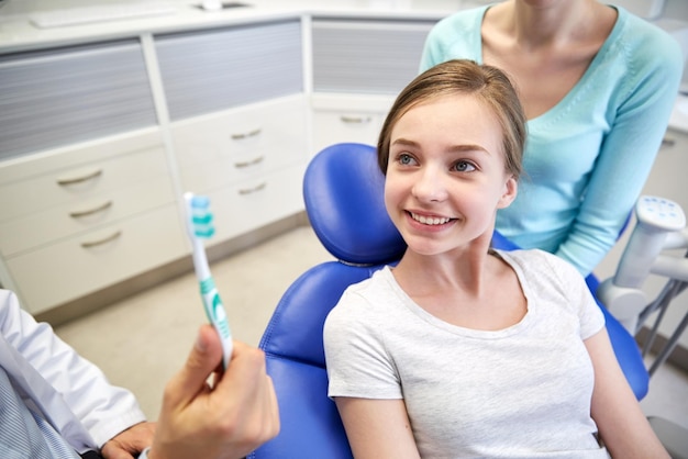 people, medicine, stomatology and health care concept - happy male dentist showing toothbrush to patient girl and her mother at dental clinic office