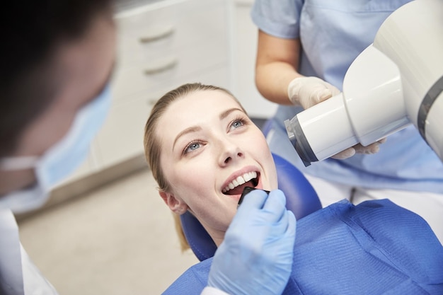 people, medicine, stomatology, technology and health care concept - close up of dentist putting intraoral shield to female patient mouth and assistant directing x-ray machine at dental clinic