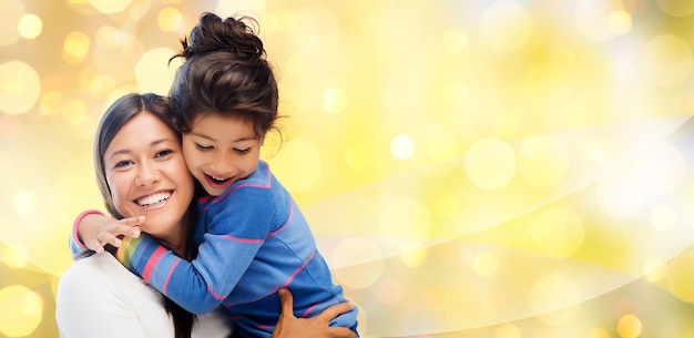 people, motherhood, family, holidays and adoption concept - happy mother and daughter hugging over yellow lights background